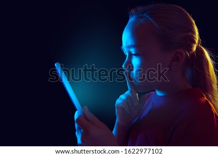 Serious. Caucasian girl's portrait isolated on dark studio background in neon light. Beautiful female model using tablet. Concept of human emotions, facial expression, sales, ad, modern tech, gadgets.