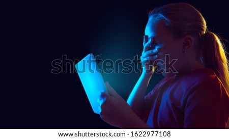 Shocked, scared. Caucasian girl's portrait on dark background in neon light. Beautiful female model using tablet. Concept of human emotions, facial expression, sales, ad, modern tech, gadgets. Flyer.