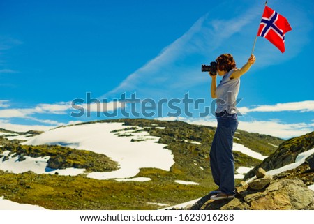 Tourist woman holding norwegian flag and taking photo with camera, snowy mountains landscape, summertime. National tourist route Aurlandsfjellet.
