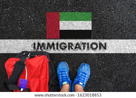 Man in shoes with bag standing next to line with word IMMIGRATION and flag of United Arab Emirates on asphalt road