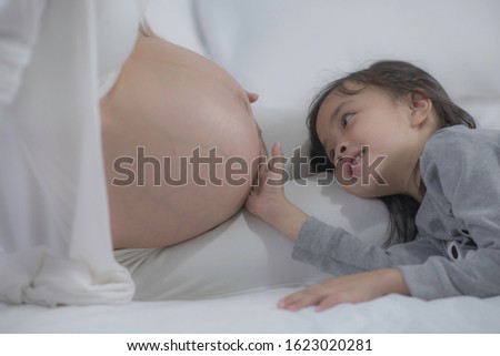 Asia little girl touching her pregnant mom belly. Expecting mother sitting   with her daughter sleep on  bed having fun and feeling baby pushes,soft picture tone.