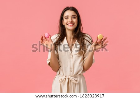 Sweets, happiness and women concept. Cheerful and cute european girl smiling happily, holding two macarons and grinning delighted, sharing dessert with friend, standing pink background