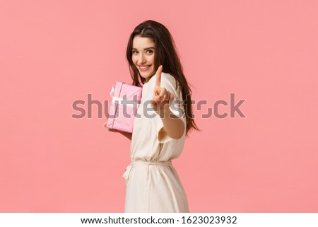Romance, relationship and holidays concept. Lovely alluring girlfriend prepared special gift for someone, show finger asking wait, hiding gift box and smiling, have surprise present, pink background