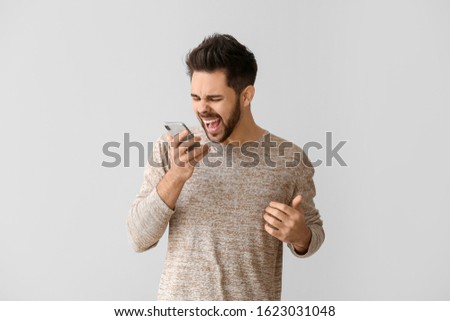 Screaming young man with mobile phone on light background