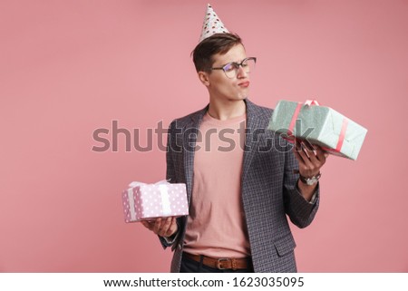 Image of a young thinking serious guy in glasses holding birthday present gift boxes isolated over pink wall background.