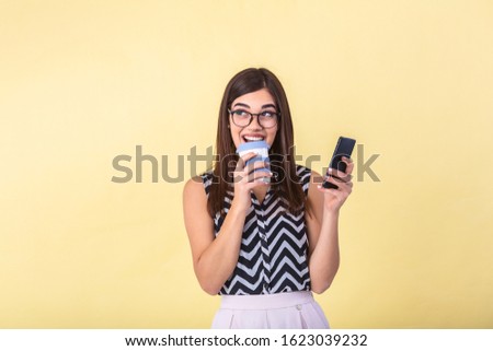 Indoor picture of young good-looking European woman isolated on peach background holding coffe to go and phone in hand and smiling while browsing or reading message