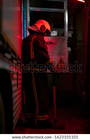 Full growth firefighter man with hammer on his shoulder standing near fire engine