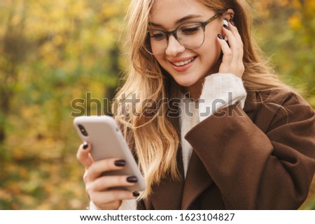 Beautiful young cheerful woman wearing coat walking in the autumn park, wearing wireless earphones, using mobile phone