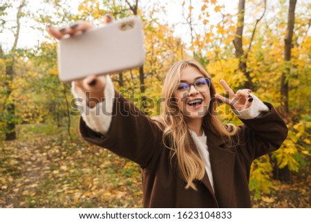 Beautiful young cheerful woman wearing coat walking in the autumn park, taking a selfie