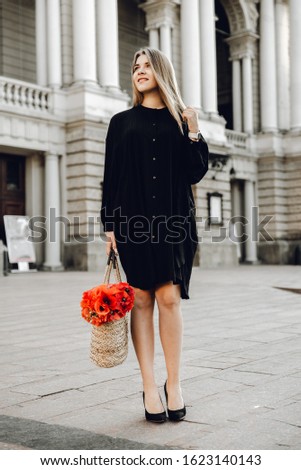 A beautiful young woman in a black dress with blond hair walks around the city with a bag with red flowers; photo of happy smiling woman standing on the square on sunny summer or spring day outside