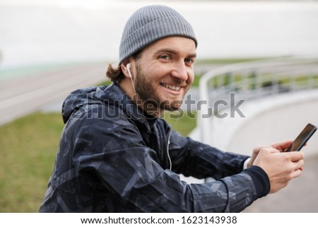 Motivated smiling young fit sportsman listening to music with earphones while standing at the stadium, using mobile phone, leaning on the rail