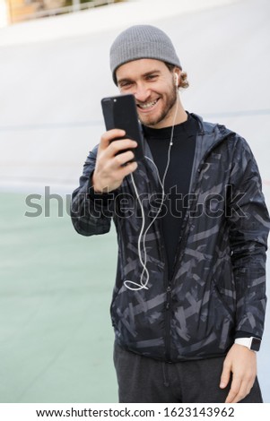 Motivated smiling young fit sportsman listening to music with earphones while standing at the stadium, taking a selfie with mobile phone