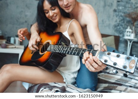 Young mixed race couple boyfriend teaching girlfriend playing guitar sitting on bed at home together musical instrument close-up blurred background smiling happy