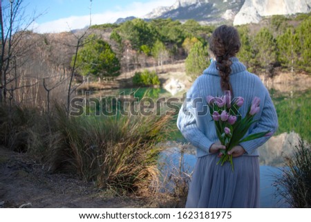 Woman posing outdoor in the nature with bouquet of flowers and enjoy the gorgeous view of lake, hills and sun. female hands holding pink tulips. Happy model dressed blue sweater. 