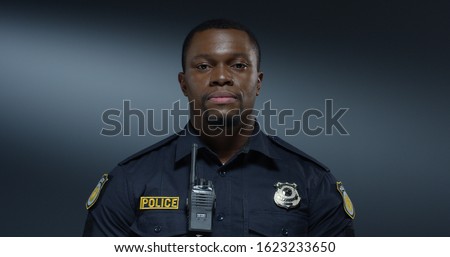 Portrait shot of the handsome young African American policeman in the uniform and walkie-talkie looking straight to the camera with serious face. Close up.