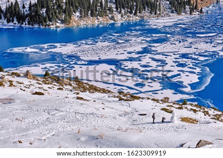 Tourists take pictures of a snowman on a clear sunny day on the background of mountain lake covered with ice in the winter season; Big Almaty lake in Kazakhstan