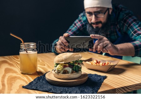 chef taking a photo with the phone of his vegan burger with lettuce and sauce, fries with ketchup and healthy drink on a wooden table, technology and gourmet lifestyle concept, selective focus