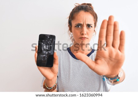 Young redhead woman holding broken smartphone over isolated background with open hand doing stop sign with serious and confident expression, defense gesture