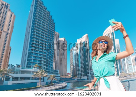 Happy asian girl taking selfie photo on a smartphone while walking on a promenade in Dubai Marina district. Travel and lifestyle in United Arab Emirates