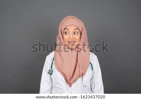 Oops! Portrait of muslim doctor female clenches teeth and looks confusedly aside, realizes her bad mistake, poses against copy area for your advertising or promotional background.