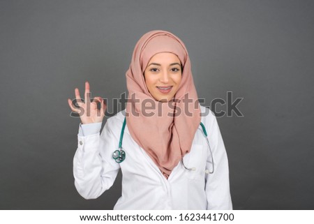 Glad attractive muslim doctor woman shows ok sign with hand as expresses approval, has cheerful expression being optimistic. Standing against gray wall.