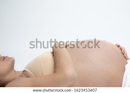 Close up of the pregnant woman lying on the ground and holds her hands on her swollen belly, isolated over white background, studio photo