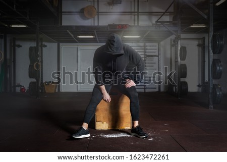 Young fit man in black sweatsuit with hood on his head sitting on the jump box in the gym taking a rest after hard core cross workout training Royalty-Free Stock Photo #1623472261