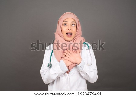 Happy doctor muslim woman standing and smiling isolated on gray studio background has hands on chest near heart. Young emotional woman. Human emotions, facial expression concept. Front view.