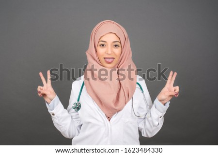 Indoor portrait of young caucasian muslim doctor female isolated on gray background with optimistic smile, showing peace or victory gesture with both hands, looking friendly. V sign.