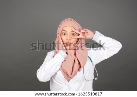 Leisure lifestyle people person celebrate flirt coquettish concept. Beautiful doctor woman wearing medical uniform showing v-sign near eyes wearing casual clothes standing against gray wall.