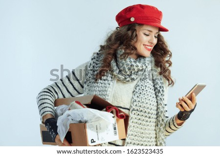 smiling trendy female with long brunette hair in sweater, scarf and red hat holding opened parcel and using smartphone app isolated on winter light blue background.
