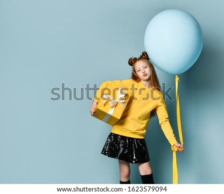 Red-headed teen kid in yellow sweatshirt, black skirt, knee-highs, boots. Kissing you, holding balloon and golden gift box, posing against blue background. Hipster style, fashion, holiday. Full length