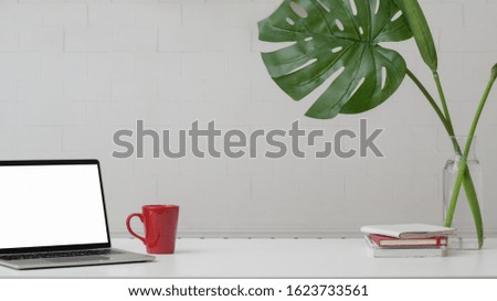 Cropped shot of stylish home office with copy space, blank screen laptop, decorations and coffee cup on white table with white wall background  