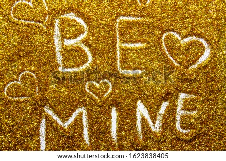Drawing a words  be mine on the golden sand glitter. A words be mine against a gold texture on the glitter background. A Shiny gold glitter on the background