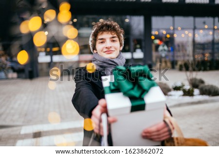 Happy adult hipster teenager man walking on street over shopping center on black friday sales, carrying shopping bags and showing gift box with green bow looking at camera with smile outdoor in winter