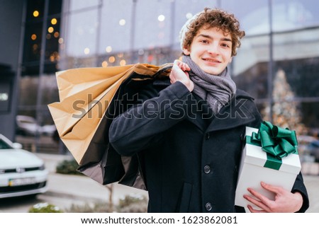 Man holding a heap of gift boxes. Boyfriend caring an impressive stack of holiday presents packed with nice ribbons to give for special occasion or event. Close up outdoor portrait of curly guy.