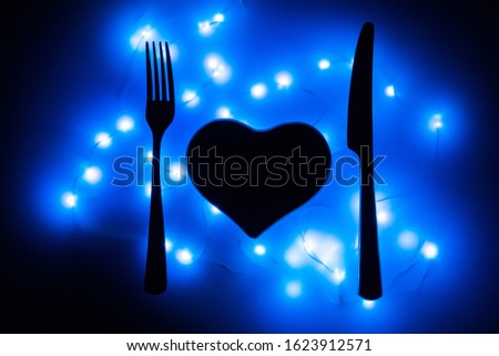 Black heart, fork and knife in the dark surrounded by blue garlands. The heart is served for dinner. Table setting for Valentine's day. Valentine's day February 14. Love is presented as food.
