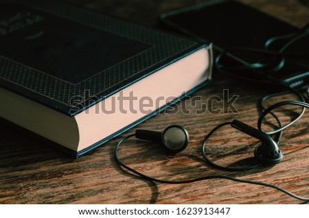 Big blue book, earphones and smartphone on a wooden table. Closed hardcover book. Side view. Eye level shooting. Selective focus. Without people. Close-up. Tinted photo