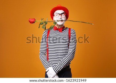 Mime with white face in red hat and striped T-shirt holding rose in his mouth on blank orange background