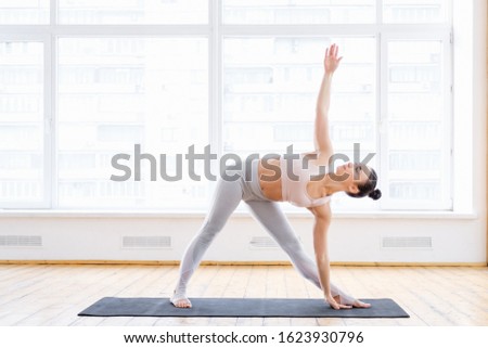 Cute slim Caucasian fitness model doing utthita trikonasana exercise, extended triangle pose in spacious gym with large windows. Concept strong healthy back and good stretching. Space for advertising