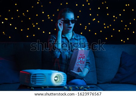 girl in 3D glasses with popcorn on a sofa at home watching movies through a projector