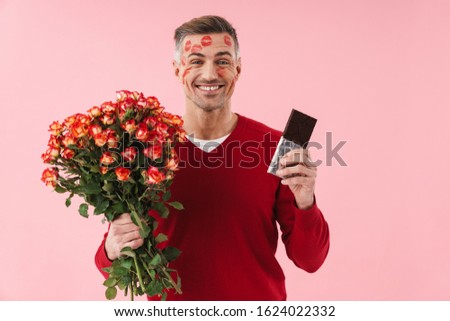 Portrait of handsome caucasian man with kiss marks at his face holding flowers and chocolate bar isolated over pink background