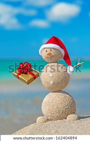 Smiley sandy snowman at beach in christmas hat with golden gift. Holiday concept for New Years Cards.