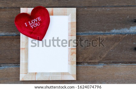 Square picture frame with a red heart resting on a frame. In the heart there is a message that I love you. All of this is laid on an old wooden plate.