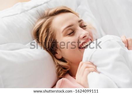 Image of a pretty happy smiling optimistic young woman indoors at home lies in bed under blanket.