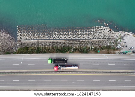 Top down view of trucks driving on a coastal highway that is protected with the concrete wave breaker & the green seawater shining under the summer sun, in Bali District, New Taipei City, Taiwan, Asia