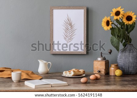 Stylish and sunny interior of kitchen space with wooden table, brown mock up photo frame, breakfast and sunflowers. Scandinavian home decor with kitchen accessories. Template. Gray color concept