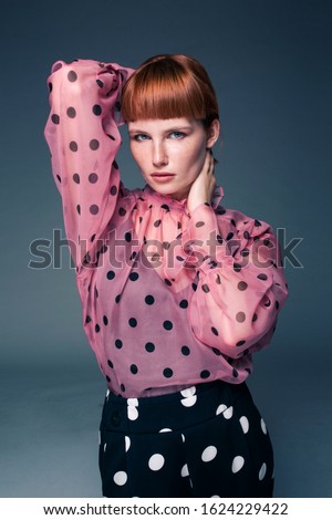 Young fashionable red hair woman wearing nice pink blouse with big black dots. Portrait of happy smiling girl in stylish glamorous clothes. Autumn winter spring fashion photo on a grey background.