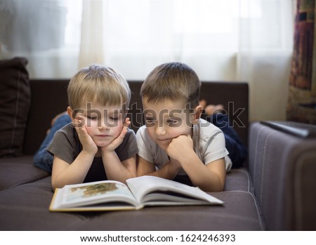 Two cute boys in T-shirts, brothers of the same age 3-4 years old, lie on their stomachs on a brown sofa, propping  hands on their heads, with interest, carefully examine the book in front of them
