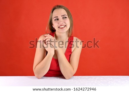Universal concept female portrait on a red background. Horizontal photo of a pretty smiling girl with long hair and great makeup sitting at a white table in the studio.